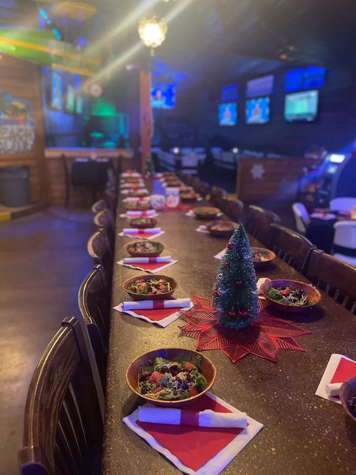 A long table set for dinner with red napkins and a small lit Christmas tree down the centre